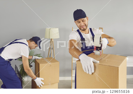 Two professional young male movers securing cardboard boxes with adhesive tape in new home. Two professional young male movers securing cardboard boxes with adhesive tape in new home. 129655816