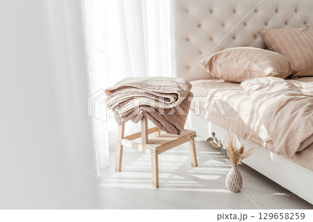 Cozy folded beige blanket on wooden stool next to a neatly made bed in morning sunlight 129658259