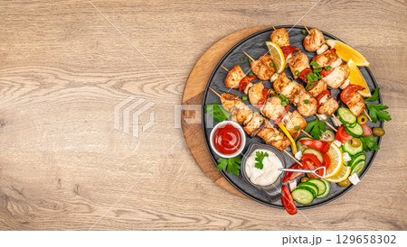 Grilled chicken skewers with fresh vegetable salad, lemon wedges, and dipping sauces on a round black platter. top view. copy space 129658302