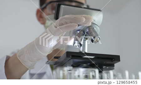 Female scientist wearing white protective gear analyzing test samples under microscope in contemporary research environment, close up of white gloved hand. Medicine and science concept Female scientist wearing white protective gear analyzing test samples under microscope in contemporary research environment, close up of white gloved hand. Medicine and science concept 129658455