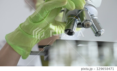 Close up of green gloved hands of a scientist adjusting a microscope in a laboratory, emphasizing the significance of scientific research and analysis. Medicine, healthcare and science concept 129658471
