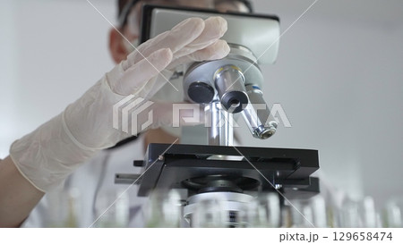 Female scientist wearing white protective gear analyzing test samples under microscope in contemporary research environment, close up of white gloved hand. Medicine and science concept Female scientist wearing white protective gear analyzing test samples under microscope in contemporary research environment, close up of white gloved hand. Medicine and science concept 129658474