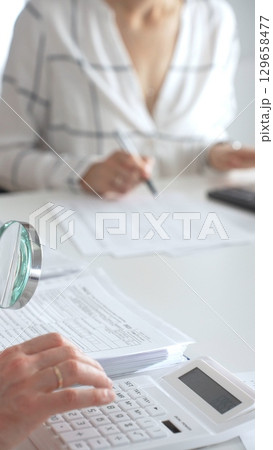Two accountants are working together, using a magnifying glass and a calculator, to audit financial reports in their brightly lit office. Audit and taxes in business 129658477