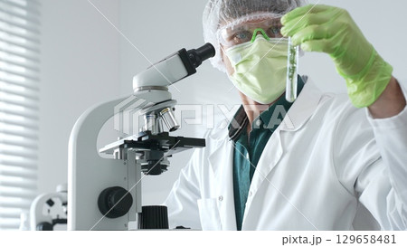 Adult male scientist wearing green protective gear looking at plant samples in test tubes and using a microscope in modern laboratory for botanical research. Medicine, healthcare and science concept Adult male scientist wearing green protective gear looking at plant samples in test tubes and using a microscope in modern laboratory for botanical research. Medicine, healthcare and science concept 129658481