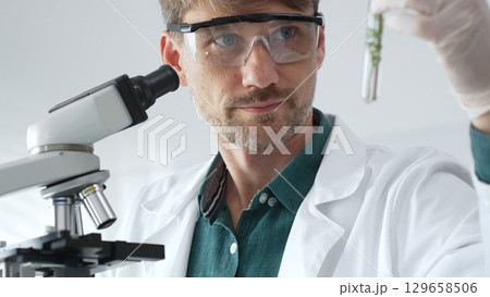 Adult male scientist in mask and gloves holding test tube with green plant sample, conducting laboratory research with a microscope. Medicine, health care and science concept Adult male scientist in mask and gloves holding test tube with green plant sample, conducting laboratory research with a microscope. Medicine, health care and science concept 129658506
