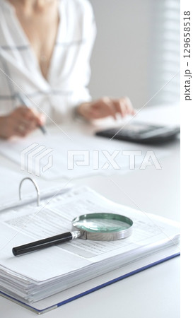 Close up of a magnifying glass on financial documents, with an accountant using a calculator and taking notes, emphasizing accounting, finance, and tax preparation. Audit and taxes in business 129658518