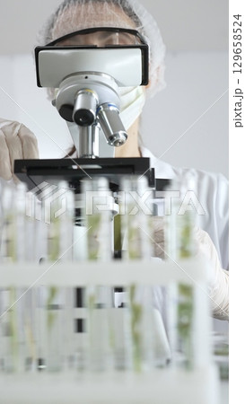 Scientist wearing green protective gear using microscope, analyzing test tubes in laboratory, conducting scientific research and development. Medicine, healthcare and science concept Scientist wearing green protective gear using microscope, analyzing test tubes in laboratory, conducting scientific research and development. Medicine, healthcare and science concept 129658524