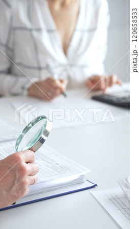 Close up of accountant hand using a magnifying glass and a calculators, to audit financial reports in brightly lit office. Audit and taxes concept 129658533
