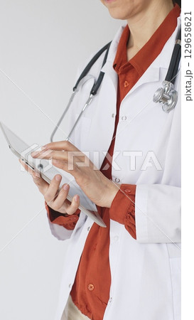 Unknown professional woman doctor, wearing white coat over a brick-red blouse and a stethoscope, is using a digital tablet, suggesting innovative healthcare practices in medicine. Health care concept Unknown professional woman doctor, wearing white coat over a brick-red blouse and a stethoscope, is using a digital tablet, suggesting innovative healthcare practices in medicine. Health care concept 129658621