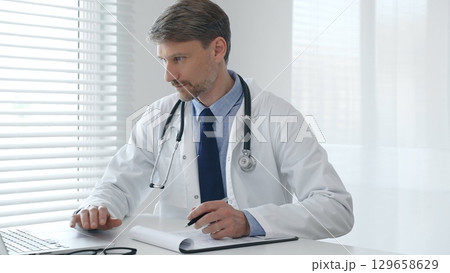 A male doctor in a lab coat with a stethoscope sits at a table in a medical office and uses a laptop while taking notes on a clipboard for patient care and treatment 129658629