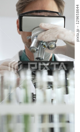 Man scientific researcher in protective gear analyzing specimens through a microscope in a sterile laboratory environment. Medicine, health care and science concept 129658644