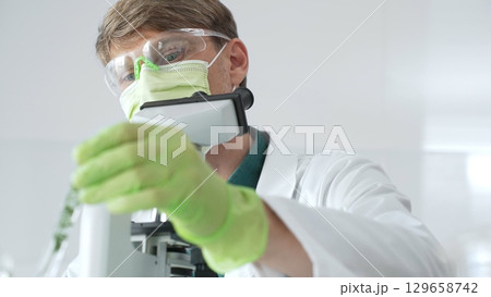 Adult male scientist wearing green mask, gloves and goggles using microscope and analyzing sample in test tube in a laboratory, developing new drugs and vaccines. Medicine and science concept 129658742