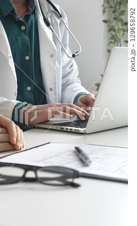 Doctor man wearing lab coat over a green shirt, is typing on a laptop and listening to a woman patient during medical consultation in clinic. Medicine and health care Doctor man wearing lab coat over a green shirt, is typing on a laptop and listening to a woman patient during medical consultation in clinic. Medicine and health care 129658792