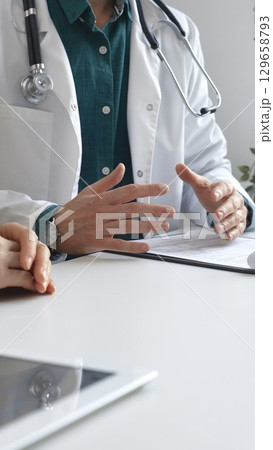 Doctor man wearing medical coat over green shirt, and stethoscope is gesturing with hands over white table while explaining diagnosis to patient. Medicine and health care concept Doctor man wearing medical coat over green shirt, and stethoscope is gesturing with hands over white table while explaining diagnosis to patient. Medicine and health care concept 129658793