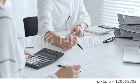 Two accountants collaborating in a well lit office, analyzing financial data and preparing reports with calculators and pens. Audit and taxes in business Two accountants collaborating in a well lit office, analyzing financial data and preparing reports with calculators and pens. Audit and taxes in business 129658802