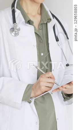 Female doctor, wearing a stethoscope and a medical coat over a green blouse, is standing and writing on a white clipboard, representing medical expertise and patient care. Medicine concept. Female doctor, wearing a stethoscope and a medical coat over a green blouse, is standing and writing on a white clipboard, representing medical expertise and patient care. Medicine concept. 129658803