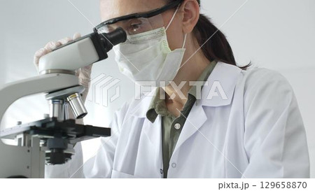 Female scientist examining microscope slide, wearing protective gear while conducting medical research in sterile laboratory environment. Medicine, healthcare and science concept Female scientist examining microscope slide, wearing protective gear while conducting medical research in sterile laboratory environment. Medicine, healthcare and science concept 129658870