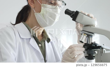 Female scientist examining microscope slide, wearing white protective gear while conducting medical research in laboratory environment. Medicine, health care and science concept Female scientist examining microscope slide, wearing white protective gear while conducting medical research in laboratory environment. Medicine, health care and science concept 129658875