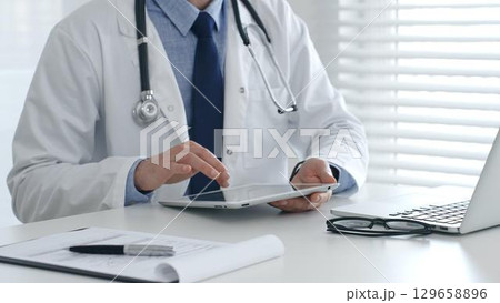 Doctor wearing lab coat and stethoscope using digital tablet while sitting at desk in medical office with laptop, eyeglasses, and medical chart nearby. Medicine and health care concept 129658896