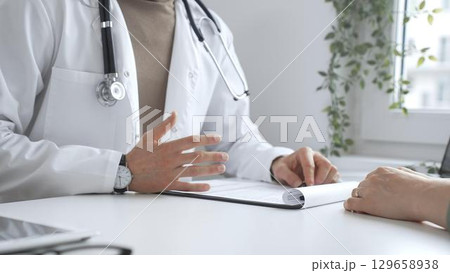 Doctor man is explaining diagnosis to female patient during a medical consultation in a medical office, gesturing with hands over clipboard and white desk. Medicine and health care concept 129658938