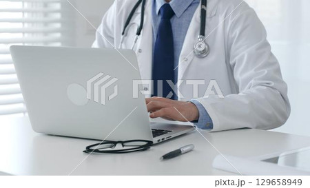 Male doctor in lab coat and stethoscope sitting at desk typing on laptop, holding glasses and pen nearby in medical office. Medicine and healthcare concept Male doctor in lab coat and stethoscope sitting at desk typing on laptop, holding glasses and pen nearby in medical office. Medicine and healthcare concept 129658949
