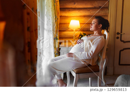 Calm and beautiful pregnant woman in white pajamas on a chair by the window. Calm and beautiful pregnant woman in white pajamas on a chair by the window. 129659813