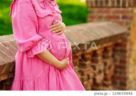 pregnant woman in pink dress hugging belly by the stone fence in the park pregnant woman in pink dress hugging belly by the stone fence in the park 129659948