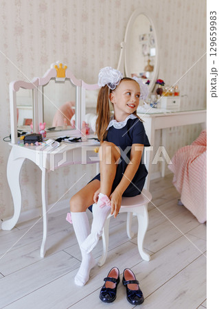 A little girl in a school uniform puts on knee socks at a dressing table.  129659983