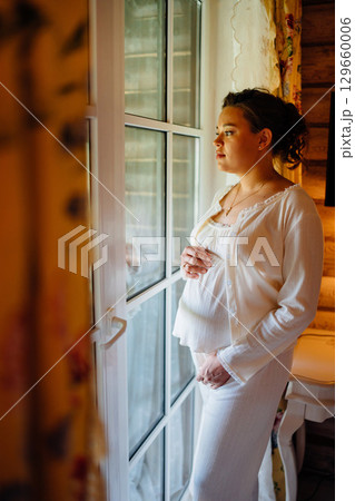 Calm and beautiful young pregnant woman in white pajamas by the window. Calm and beautiful young pregnant woman in white pajamas by the window. 129660006