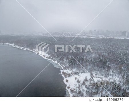 Snow-covered forest on lake shore with the first ice at sunset and the city on horizon, aerial view 129662764