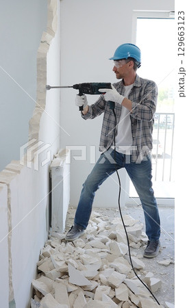 Man construction worker wearing grey checked shirt and blue hardhat is demolishing a wall with a hammer drill, creating construction debris on the surrounding floor surface. Renovation and repair 129663123