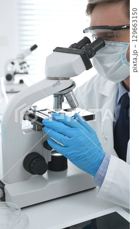 Man scientist wearing protective googles and blue gloves, is using microscope analyzing samples in modern laboratory doing scientific research. Medicine and science concept Man scientist wearing protective googles and blue gloves, is using microscope analyzing samples in modern laboratory doing scientific research. Medicine and science concept 129663150