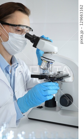 Female scientist wearing mask and blue gloves using microscope doing scientific research in modern laboratory with colleague working in background. Medicine, healthcare and science concept 129663162