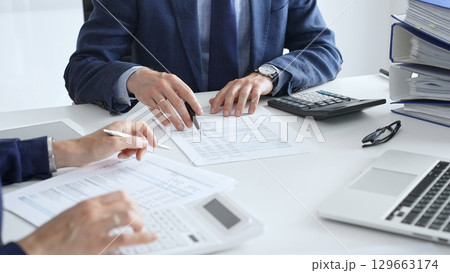 Accountants analyzing financial documents, calculating numbers together at shared workspace with laptop and office supplies surrounding them. Audit and taxes in business Accountants analyzing financial documents, calculating numbers together at shared workspace with laptop and office supplies surrounding them. Audit and taxes in business 129663174