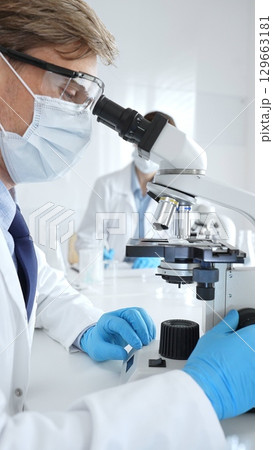 Masked male scientist analyzing sample under microscope, colleague researching nearby in sterile medical laboratory setting. Medicine, healthcare and science concept 129663181