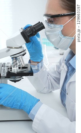 Female scientist wearing blue protective gear analyzing test samples under microscope in laboratory research environment. Medicine, healthcare and science concept Female scientist wearing blue protective gear analyzing test samples under microscope in laboratory research environment. Medicine, healthcare and science concept 129663197