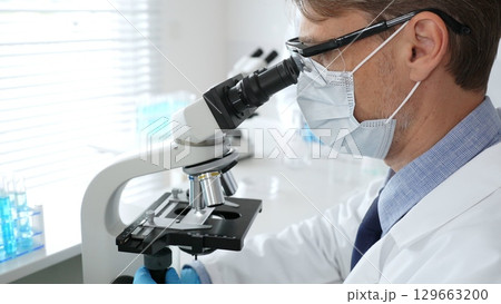 Masked scientist analyzing sample under microscope, colleague researching nearby in sterile medical laboratory setting. Medicine, healthcare and science concept 129663200