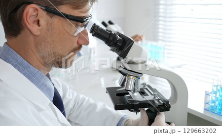 Medical scientist wearing gloves and protective glasses examining samples under a microscope in a bright laboratory filled with test tubes. Medicine, healthcare and science concept Medical scientist wearing gloves and protective glasses examining samples under a microscope in a bright laboratory filled with test tubes. Medicine, healthcare and science concept 129663202