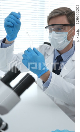 Masked male scientist in lab coat, protective glasses and blue gloves is adding reagent to test tube with microscopes and test tube rack in modern laboratory. Medicine, healthcare and science concept Masked male scientist in lab coat, protective glasses and blue gloves is adding reagent to test tube with microscopes and test tube rack in modern laboratory. Medicine, healthcare and science concept 129663203