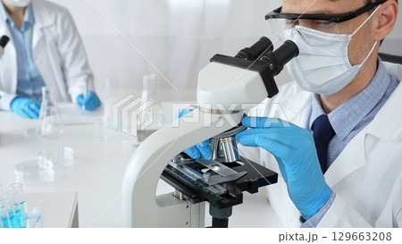 Masked scientist analyzing sample under microscope, colleague researching nearby in sterile medical laboratory setting. Medicine, healthcare and science concept 129663208