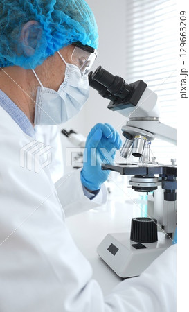 Senior scientist wearing a protective mask, cap, and gloves, examining samples under a microscope in a modern laboratory. Medicine, healthcare and science concept 129663209