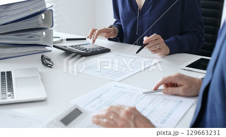 Female financial analysts with colleague examining documents, using magnifying glass and calculator while studying corporate data at workplace. Audit and taxes in business 129663231