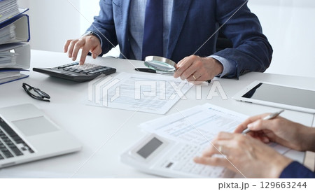 Male financial analysts with female colleague examining documents, using magnifying glass and calculator at workplace in office. Audit and taxes in business 129663244