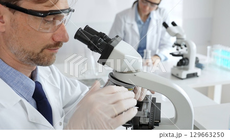 Researcher examining sample through microscope, wearing safety eyewear in sterile laboratory setting, surrounded by scientific glassware. Medicine, healthcare and science concept Researcher examining sample through microscope, wearing safety eyewear in sterile laboratory setting, surrounded by scientific glassware. Medicine, healthcare and science concept 129663250