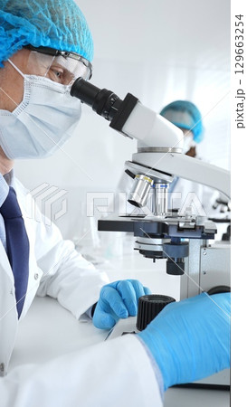 Senior male scientist wearing a protective mask, cap, and gloves, examining samples under a microscope in a modern laboratory. Medicine, healthcare and science concept 129663254