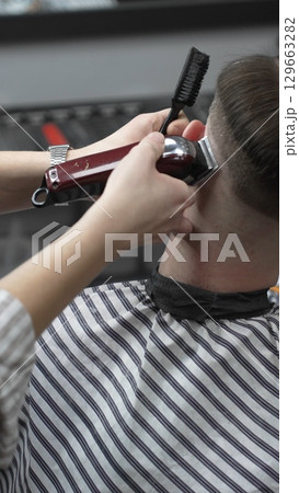 Professional barber grooming client's hair using electric clipper, delivering precise cut with skill in contemporary salon setting, close-up view Professional barber grooming client's hair using electric clipper, delivering precise cut with skill in contemporary salon setting, close-up view 129663282