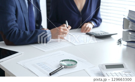 Magnifying glass and calculator over financial documents opposite financial team of male and female auditors, reviewing reports, collaborating on accounting details in office. Audit and taxes 129663300