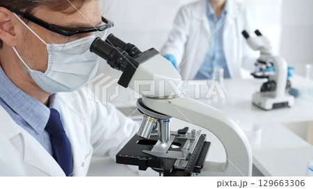 Medical researcher analyzing sample under microscope, wearing protective gear in sterile laboratory setting, performing scientific research. Medicine, healthcare and science concept 129663306