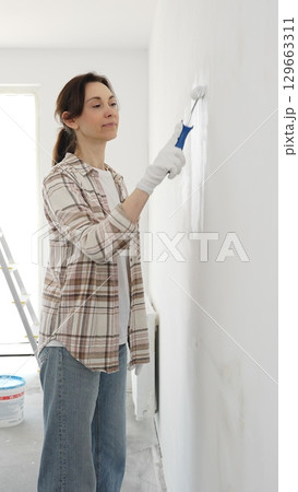 Woman wearing protective gloves rolling white paint onto wall, updating living space with fresh color and personal home renovation effort Woman wearing protective gloves rolling white paint onto wall, updating living space with fresh color and personal home renovation effort 129663311