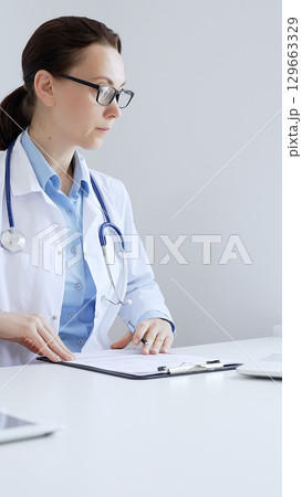 Adult doctor woman completing healthcare paperwork, clipboard positioned near laptop and stethoscope in well lit clinical workspace by window. Medicine and health care 129663329